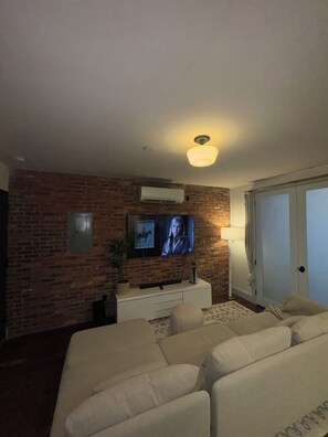Un salon avec un mur en briques, un téléviseur à écran plat fixé au-dessus d’un meuble blanc, un ventilateur de plafond et une porte-fenêtre coulissante donnant sur un balcon.