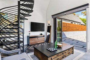 Una sala de estar moderna con una escalera espiral, un televisor de pantalla plana, una mesa de centro de madera y un ventanal corredizo que da a un área exterior.