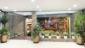 Un vestíbulo moderno con entrada de vidrio, plantas en macetas y un restaurante visible al fondo.