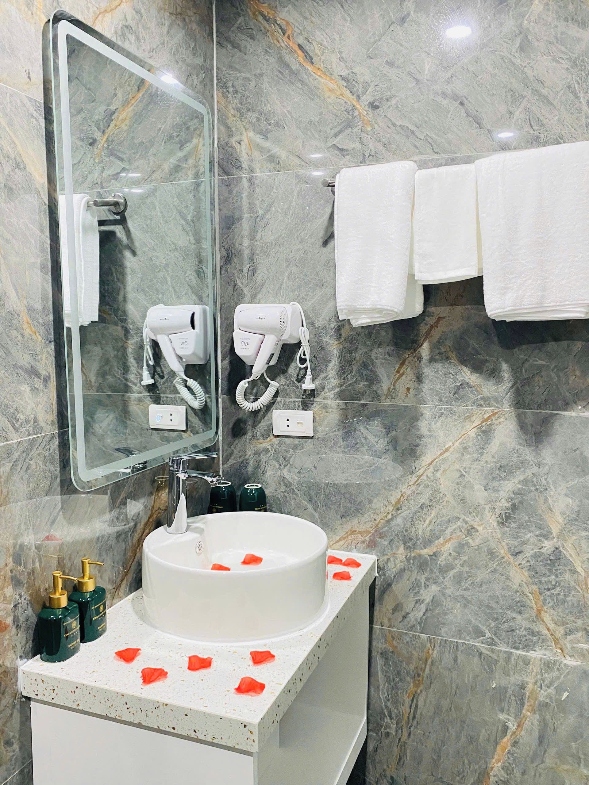 Ein Badezimmer mit Marmorwand, einem Waschbecken mit Rosenblüten, einem Föhn und Handtüchern auf einem Ablagegestell.