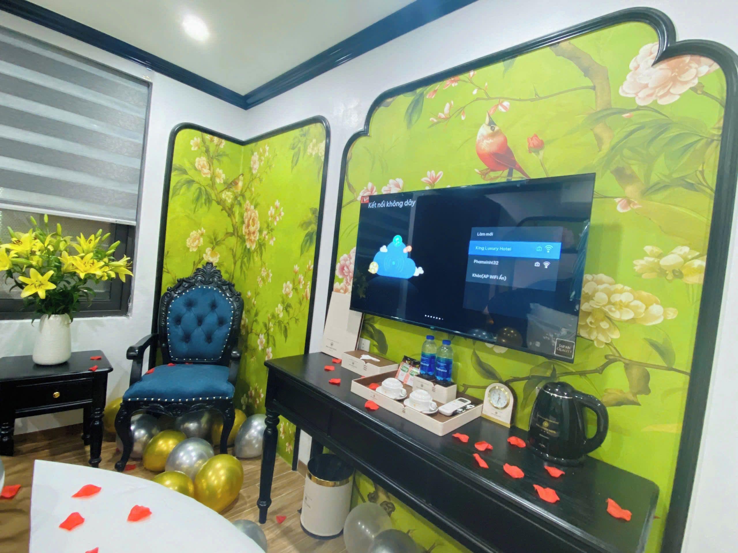 A room with a television, a blue chair, a vase with yellow flowers, and a table with various items.