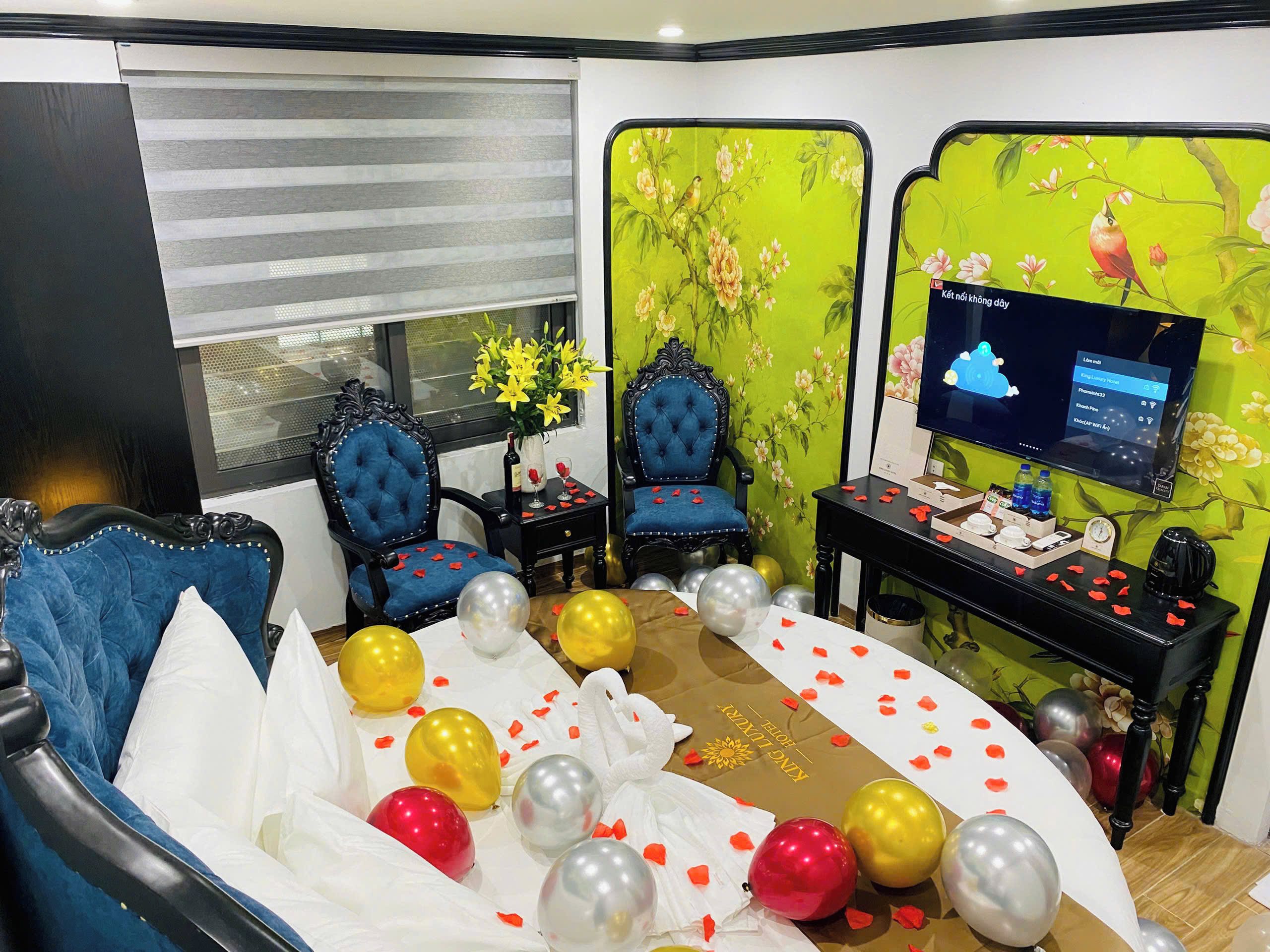 A room with a bed, two blue upholstered chairs, a small table with a vase of yellow flowers, and a television on a stand.