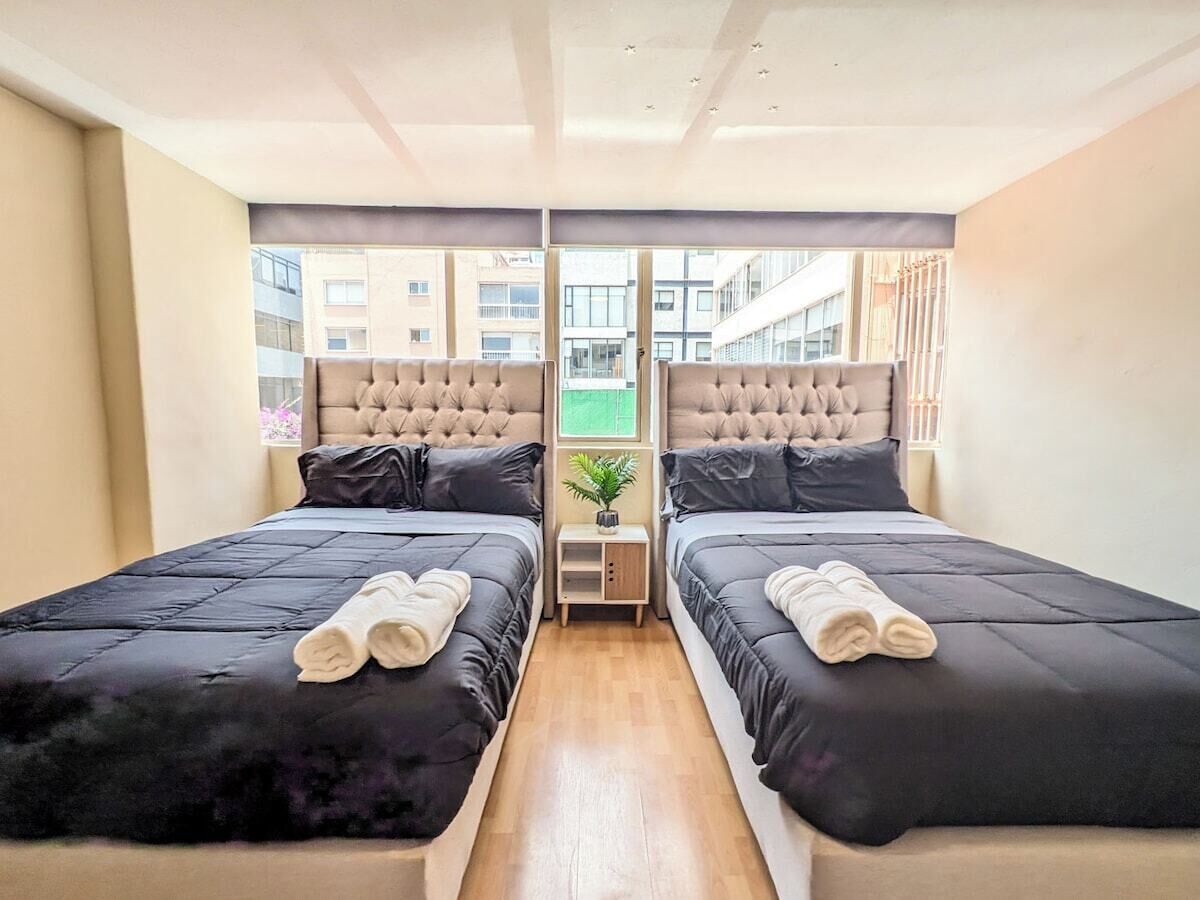 Une chambre d’hôtel avec deux lits, une petite table de chevet ornée d’une plante, et une vue sur un bâtiment à l’extérieur.