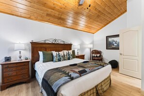 Quarto com teto de madeira, cama com colcha, duas mesas de cabeceira com abajures, uma cadeira e uma porta.