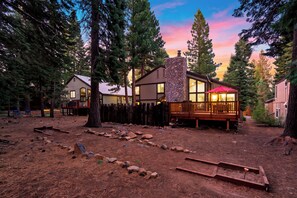 A cabin with a stone chimney, wooden deck, and a fenced yard. - Hot Tub + Big-Screen Theater + Grill Deck | Near Lifts + Trails | Gold Bear (Truckee)
