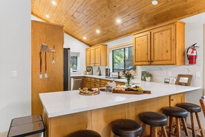 Cocina con armarios de madera, encimera blanca y una barra con taburetes.