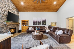 A living room with a stone fireplace, leather sofas, a coffee table, and a flat-screen TV mounted on the wall. - Hot Tub + Big-Screen Theater + Grill Deck | Near Lifts + Trails | Gold Bear (Truckee)
