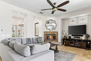 A living room with a grey sectional sofa, a fireplace, a flat-screen TV, and a ceiling fan. - Pool + Hot Tub + Game Room + Casita | Golf Course Frontage | The Back Nine (La Quinta)