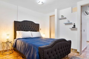 A bedroom with a tufted headboard, a bed with blue bedding, a round side table, and a small shelf with decorative items. - 2 Bedroom Apartment One Hot tub balcony (Medellin)