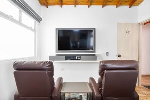 A modern living room with a flat-screen TV on a wall-mounted shelf, two leather armchairs, a small table, and a wooden door with a number. - 2 Bedroom Apartment One Hot tub balcony (Medellin)