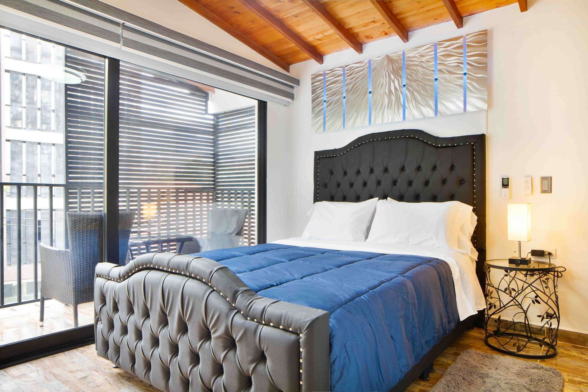 A bedroom with a large bed, a balcony with wicker chairs, and a view of buildings.