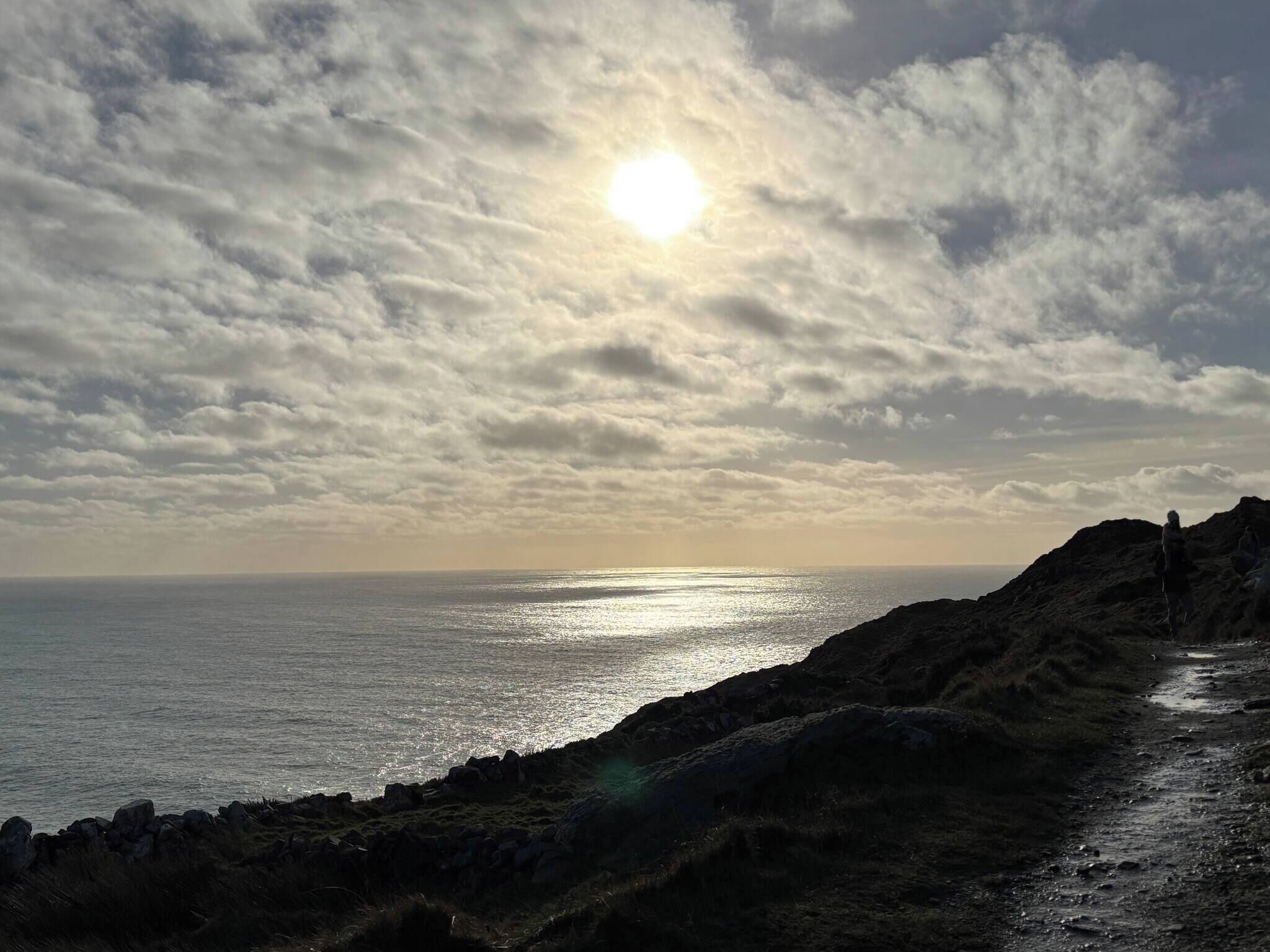 Een kustlandschap met een rotsachtig pad dat naar de zee leidt, onder een gedeeltelijk bewolkte hemel waar de zon doorbreekt.