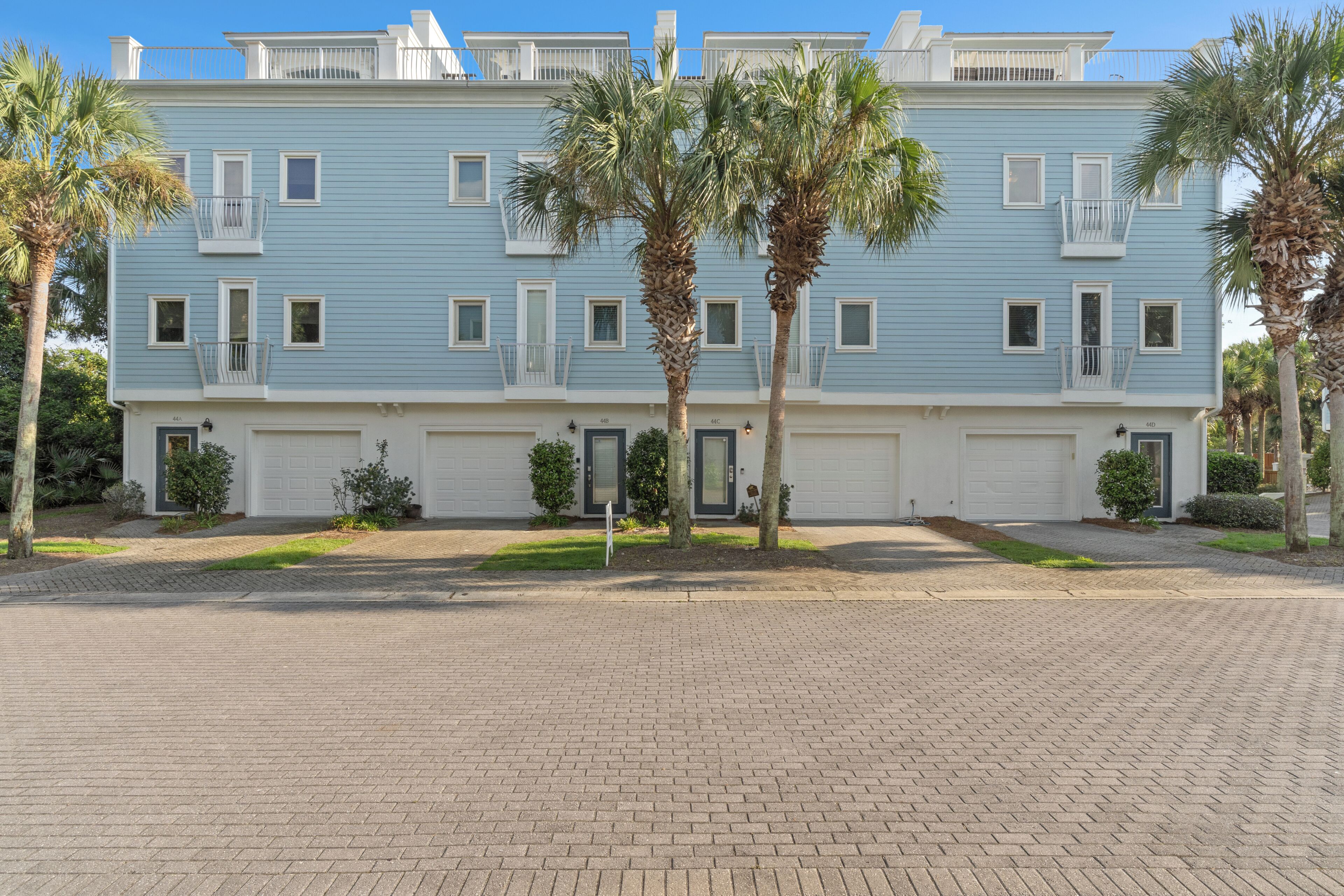 A multi-story residential building with a light blue exterior, white railings, and a brick-paved driveway.