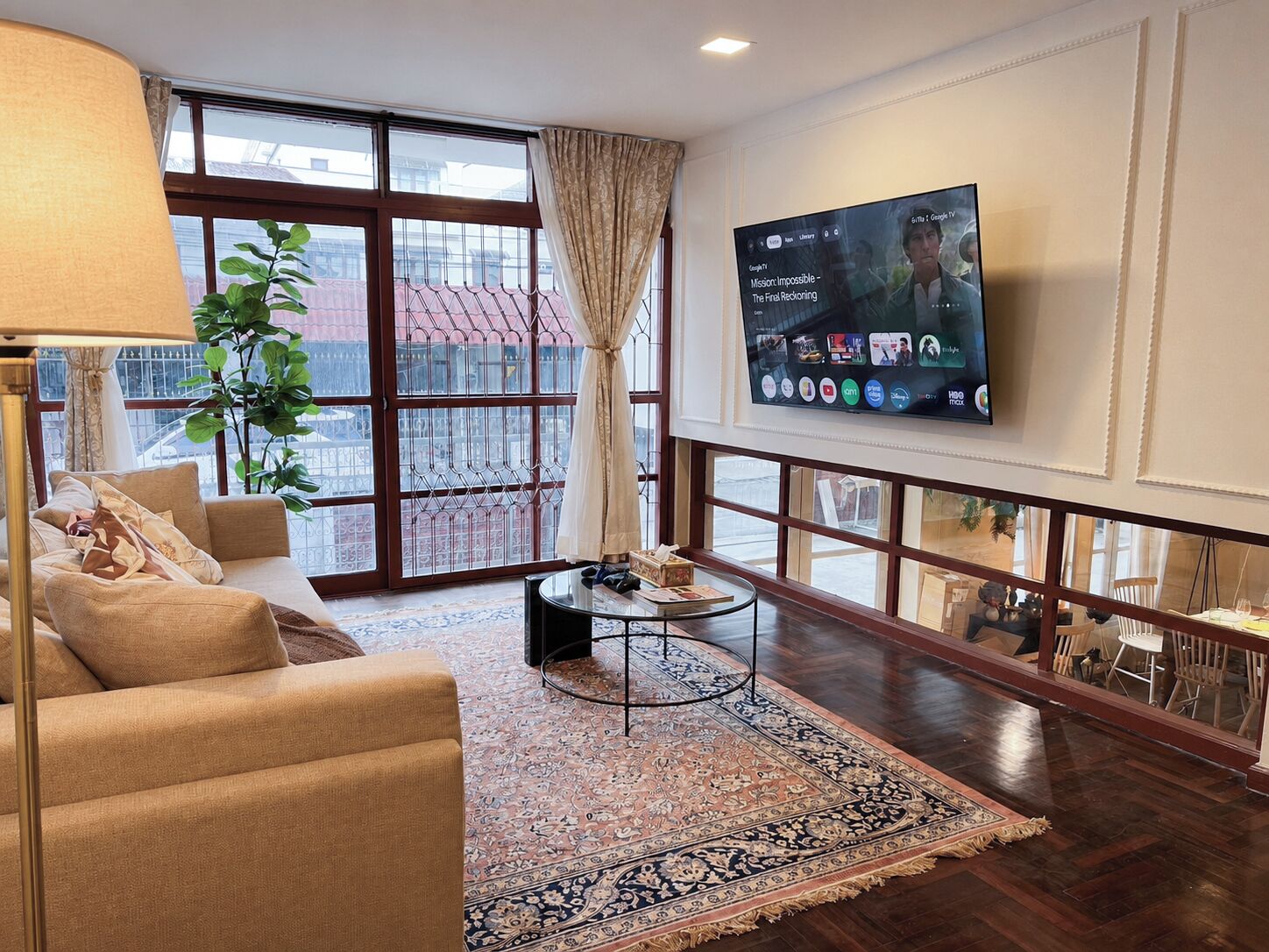 A living room with a sofa, a coffee table, a television mounted on the wall, and a large window with curtains.