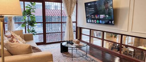 Un salon comprenant un canapé, une table basse, un téléviseur fixé au mur et une grande fenêtre avec des rideaux.