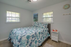 A bedroom with a bed, two windows with blinds, a wall clock, a painting, and a bedside table with a tissue box. - 1 Mi to Beach! Family-Friendly Waveland Home (Waveland)