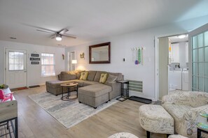 Una sala de estar con un sofá modular, una mesa de centro, un sillón estampado, un ventilador de techo, un espejo grande y un área de lavandería con lavadora.
