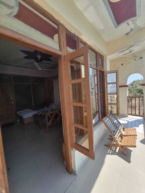 A room with a wooden door, a ceiling fan, a bed, a table with a chair, and a view of the outside.