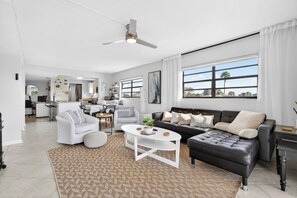 A modern living room with a white sofa, a black leather couch, and a coffee table. There is a ceiling fan, a large window with curtains, and a kitchen in the background. - Pet-Friendly Condo Near the Beach w/ Boat Slip (Treasure Island)