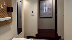 Um corredor com parede branca, uma obra de arte emoldurada, prateleira branca e um cesto de lixo.