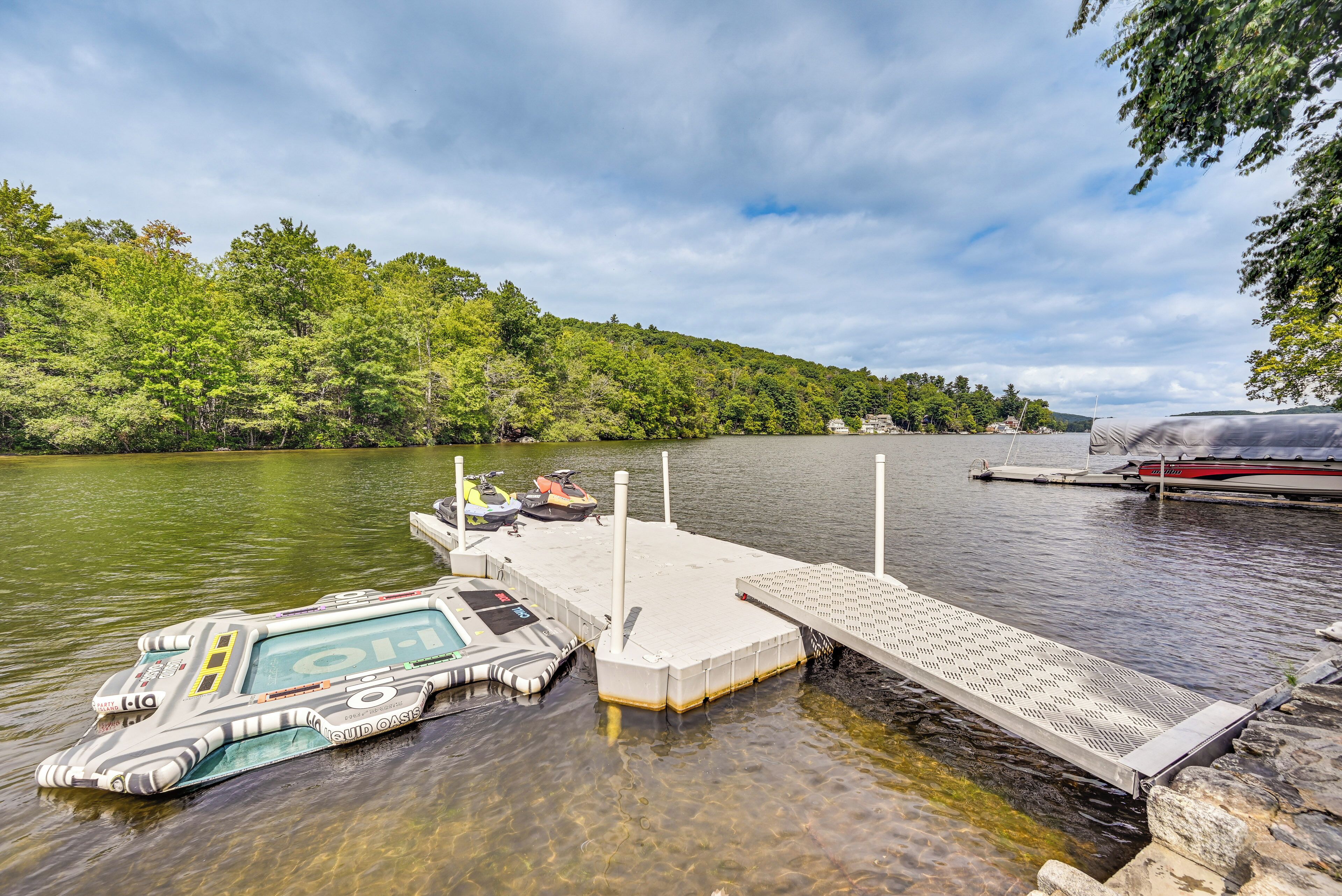 A dock with a pontoon boat and a jet ski, surrounded by a calm lake and dense greenery.