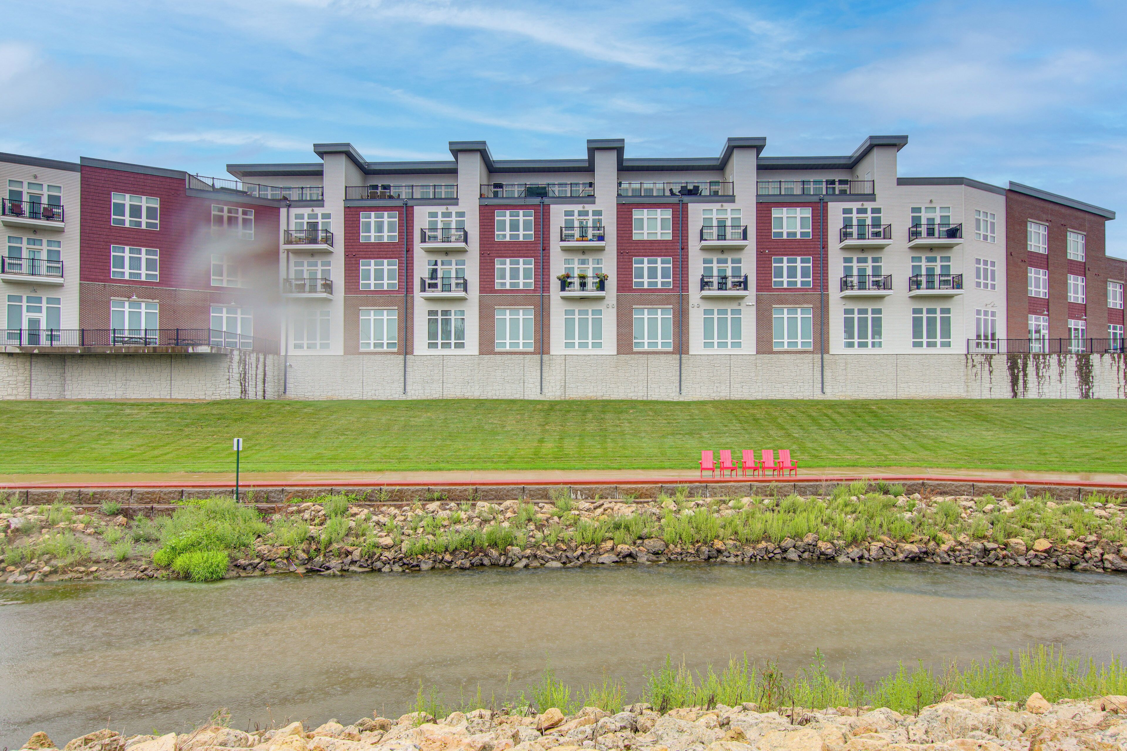 A multi-story residential building with a stone facade, a grassy area in front, and a small body of water.