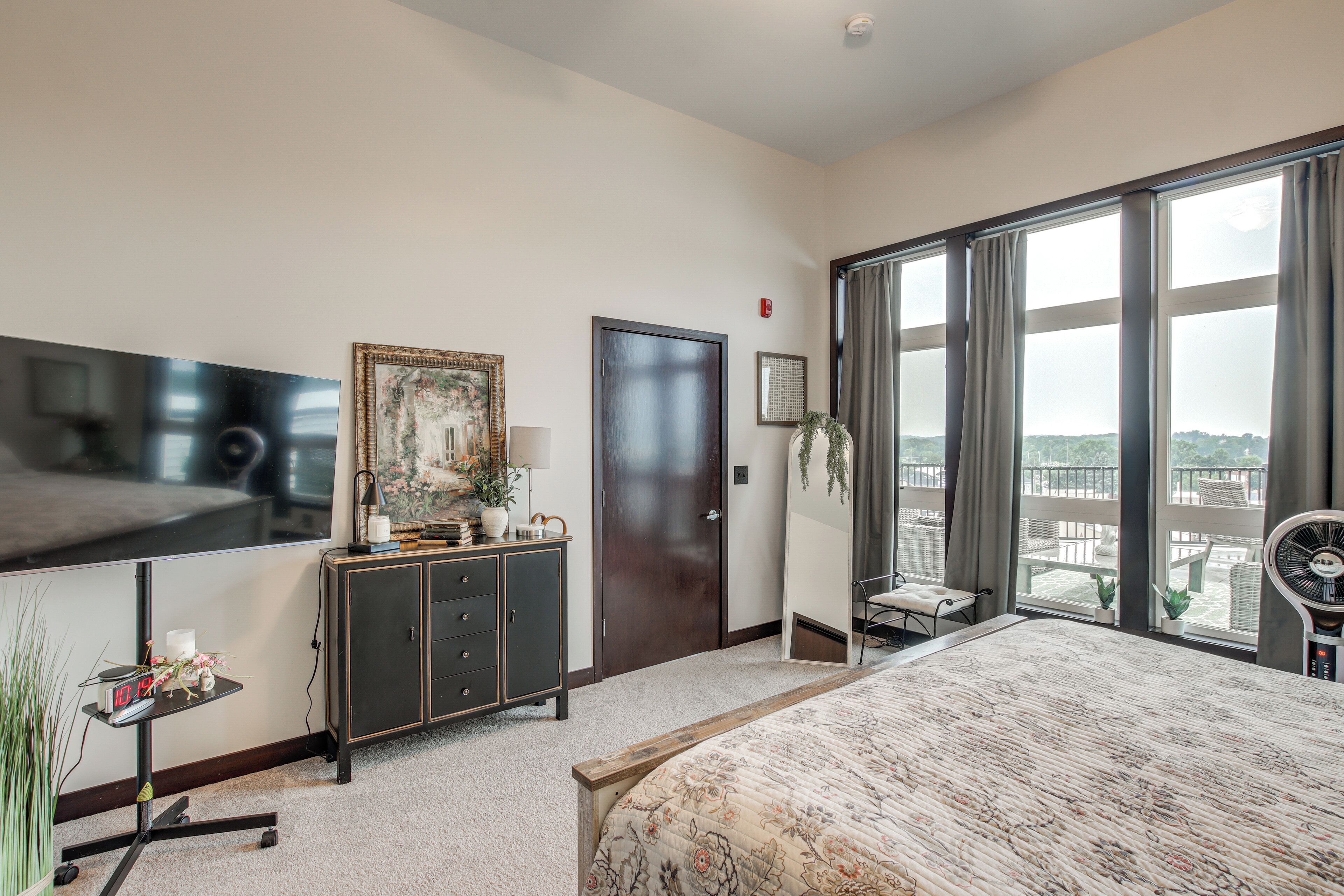 A hotel room with a large bed, a TV, a cabinet, a door, a window with a view, and a plant.