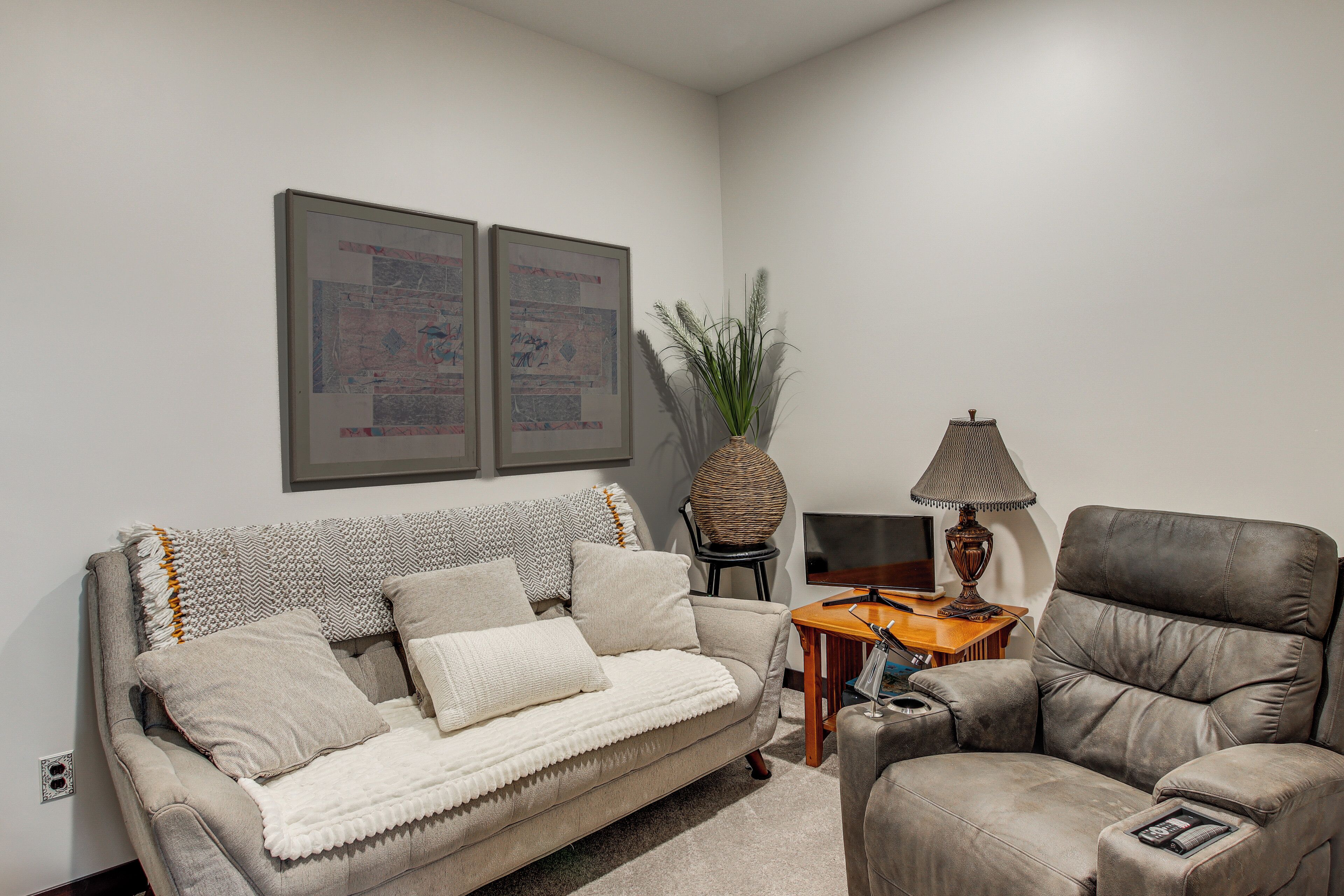 A living room with a sofa, armchair, a small table with a lamp, and two framed artworks on the wall.
