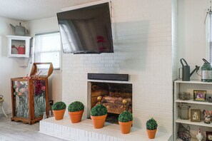 Ein Wohnzimmer mit Kamin aus weißem Mauerwerk, einem flachbildfernseher darüber und mehreren Blumentöpfen auf dem Sims.