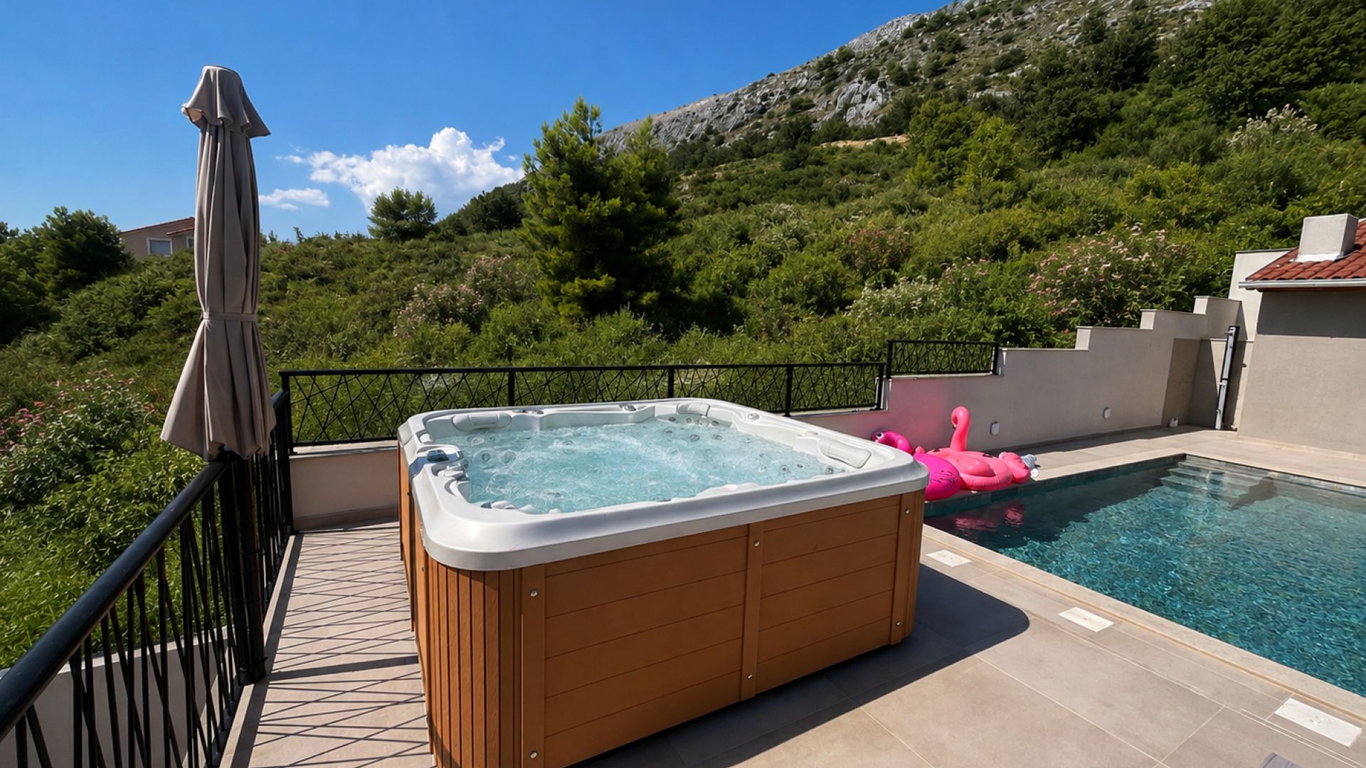 A hot tub on a balcony with a pool, surrounded by greenery and a mountain in the background.