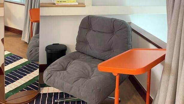 A compact room with a gray cushioned chair, a small orange table, a mirror, and a shelf with a lamp and books. - Nueng Rana 113 Unique House in Chinatown 3BR 2BA - Villa (Khet Samphanthawong)