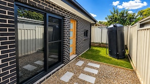A residential backyard with a paved patio, a wooden door, a black trash bin, and a white fence. - Matilda’s Urban Escape | 6-Min Station & Shops (Bradbury)