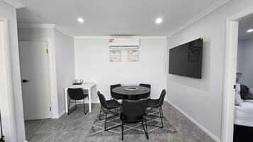 A modern room with a round table, four chairs, a white desk, a flat-screen TV mounted on the wall, and an air conditioning unit.
