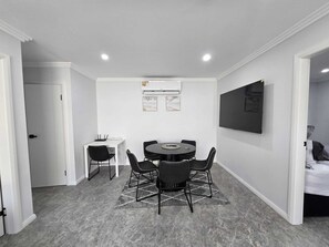 Ein modernes Zimmer mit einem runden Tisch, vier Stühlen, einem weißen Schreibtisch, einem an der Wand montierten Flachbildfernseher und einer Klimaanlage.