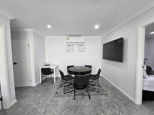 A modern room with a round table, four chairs, a white desk, a flat-screen TV mounted on the wall, and an air conditioning unit. - Matilda’s Urban Escape | 6-Min Station & Shops (Bradbury)