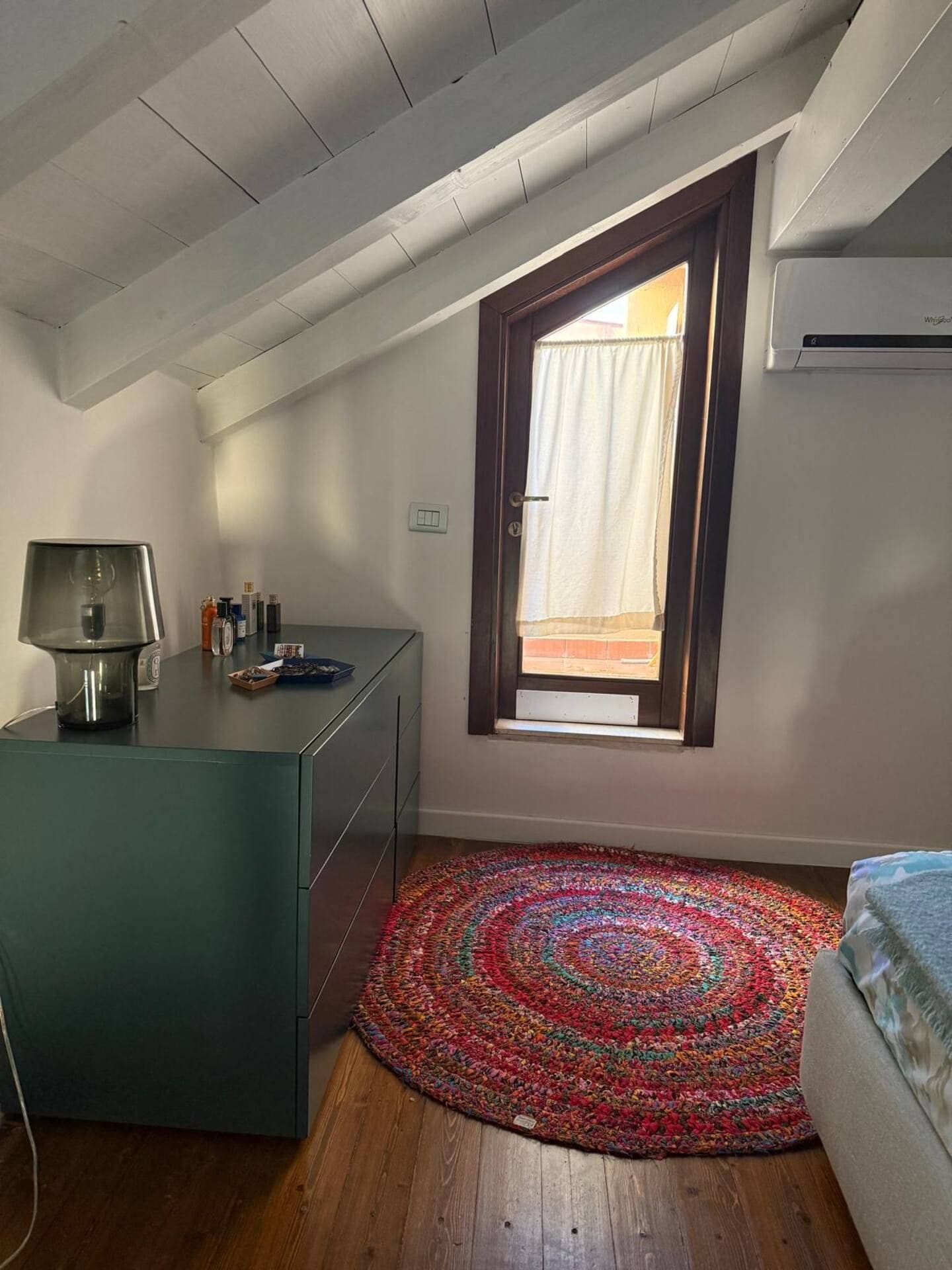 Quarto com escrivaninha, tapete colorido, janela com cortinas e aparelho de ar condicionado.