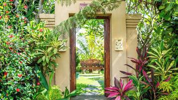 Una entrada de jardín con un arco de madera, rodeada de abundante vegetación y un sendero que lleva a un banco.
