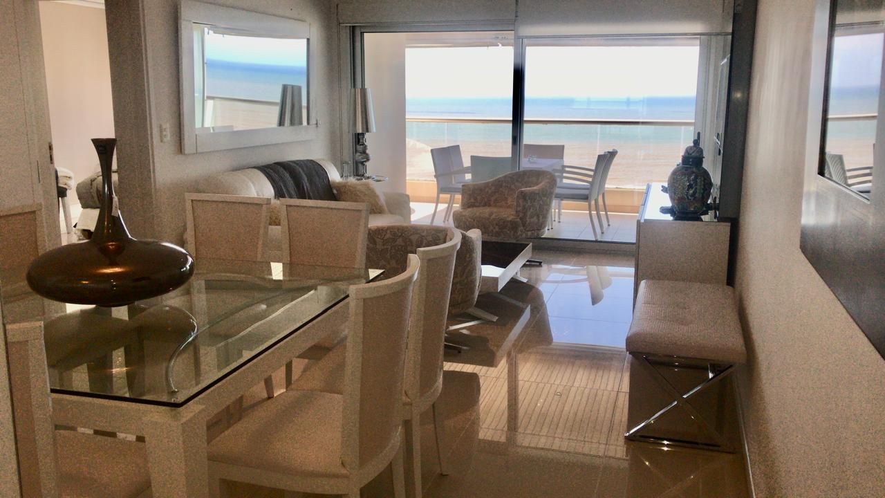 En moderne stue med et glasbord, beige sofaer og udsigt til stranden.