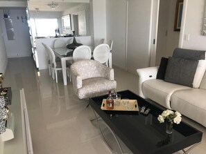 A modern living room with a glass coffee table, a sofa, and a dining area with white chairs. - Charming 2-Bedroom Apartment at Parada 6 Brava, Punta del Este (Punta del Este)