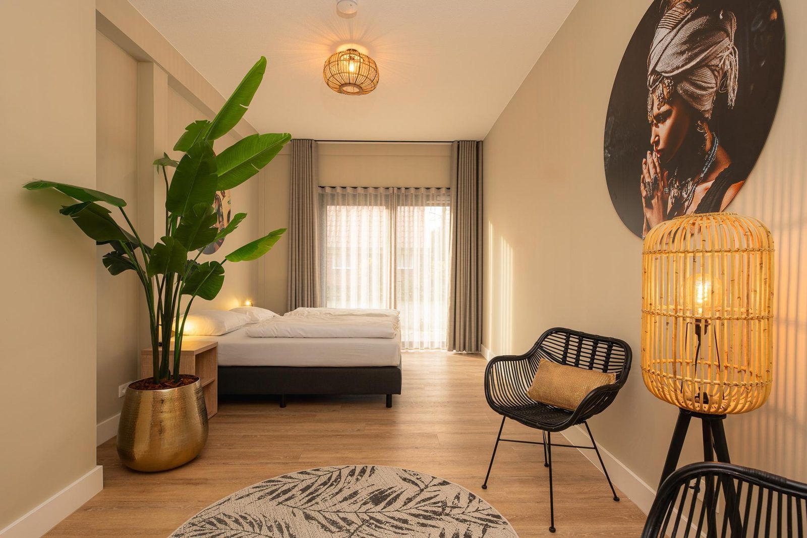 Een hotelkamer met een bed, een grote potplant, een rieten stoel, een wandlamp en een groot schilderij.