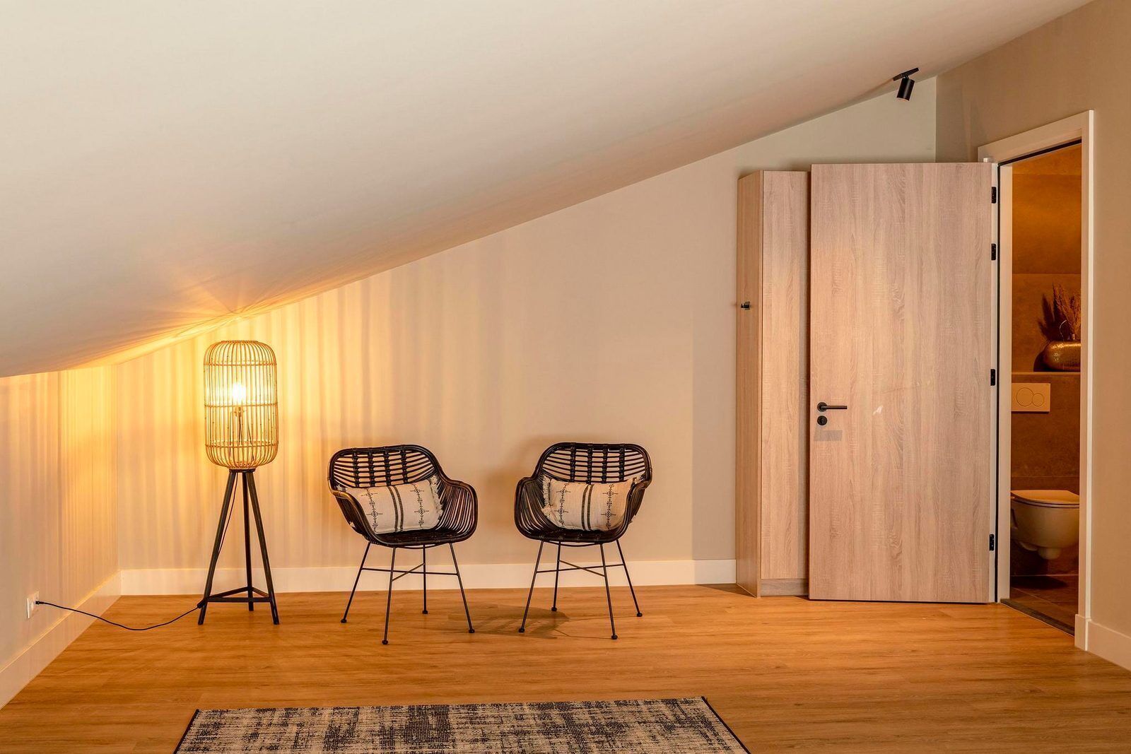 Een kamer met een schuine kap, twee stoelen met kussens, een vloerlamp en een houten deur.