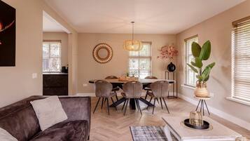 Een moderne woonkamer met een donkerbruine bank, een eettafel met stoelen, een salontafel en een groot raam met zonwering.