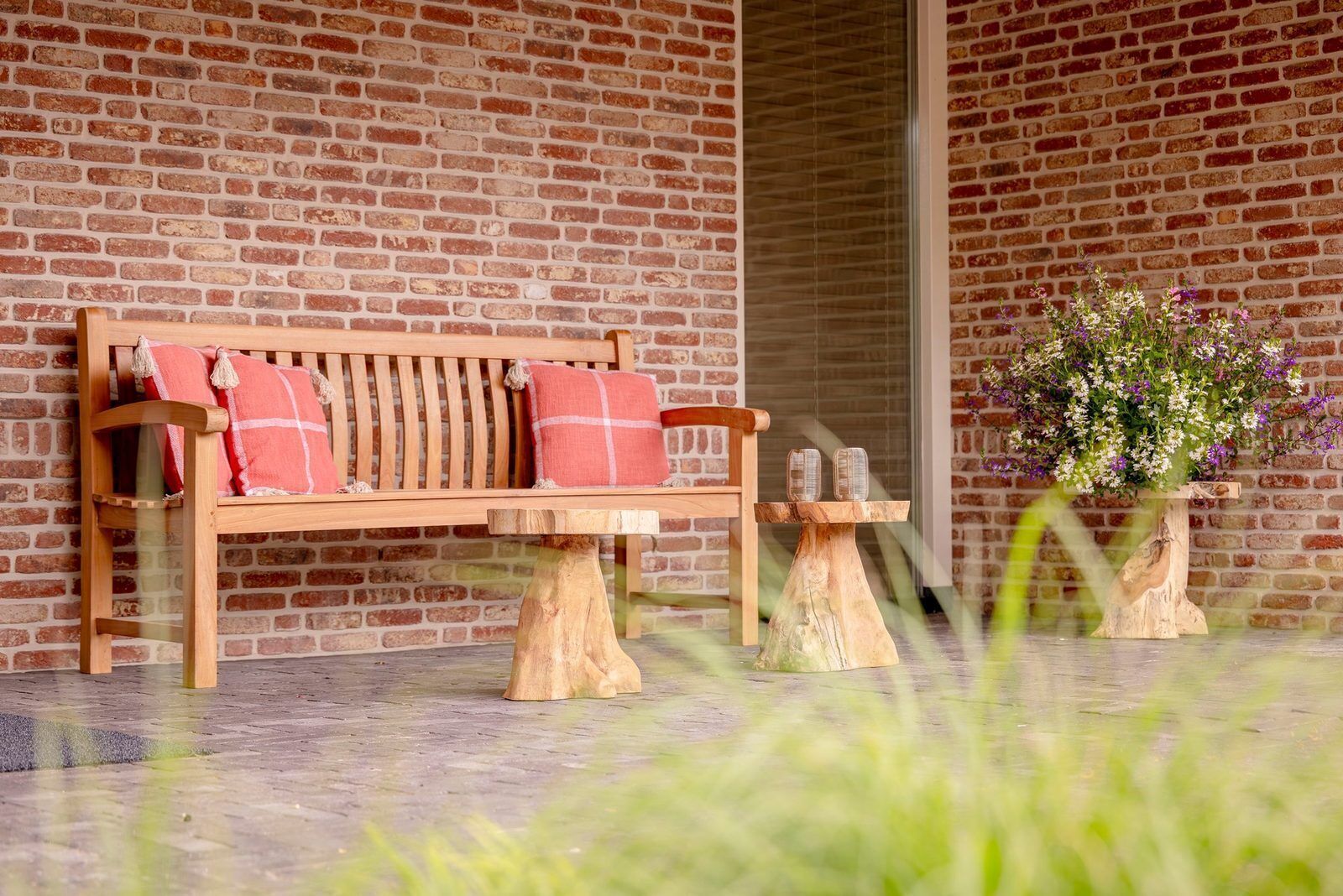 Een houten bank met een geruit kussen, een rode bakstenen muur op de achtergrond, een klein houten tafeltje met twee glazen objecten en een bloempot met witte en paarse bloemen.