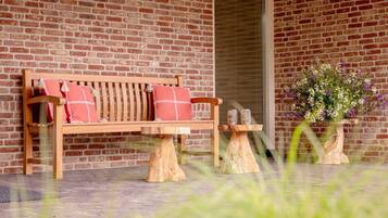 Een houten bank met een geruit kussen, een rode bakstenen muur op de achtergrond, een klein houten tafeltje met twee glazen objecten en een bloempot met witte en paarse bloemen.