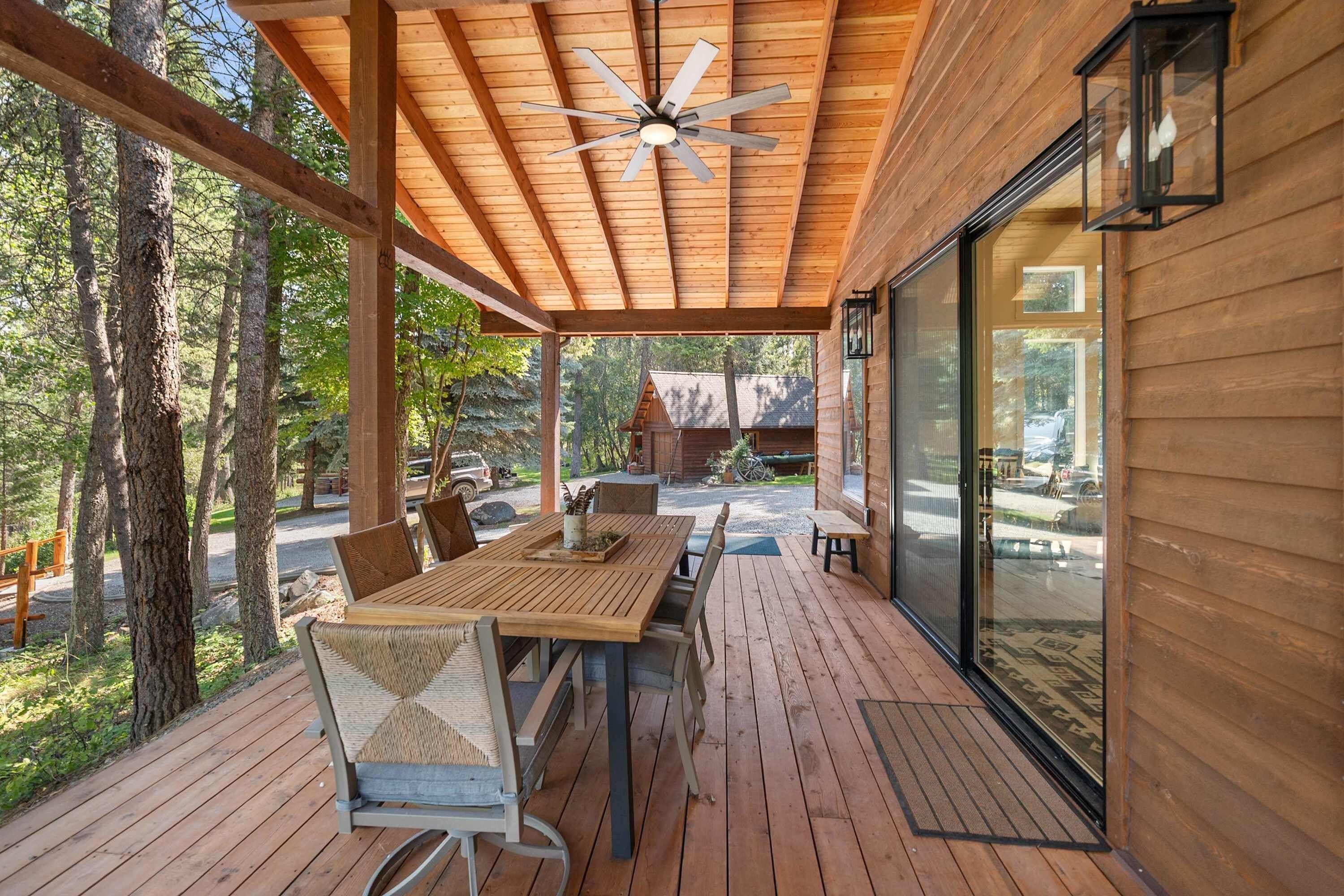 A covered outdoor seating area with a wooden table and chairs, a ceiling fan, and a view of the surrounding forest.