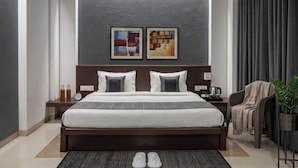 Quarto de hotel moderno com uma cama grande, duas mesas de cabeceira, um relógio e um vaso com planta.