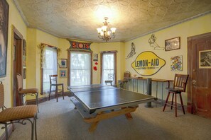 A room with a ping pong table, chairs, a chandelier, and various wall decorations including a 'SLOTS' sign and a 'LEMON-AID Customer Service' sign. - Downtown Spacious 5BR w/ Game Room (Ellicottville)