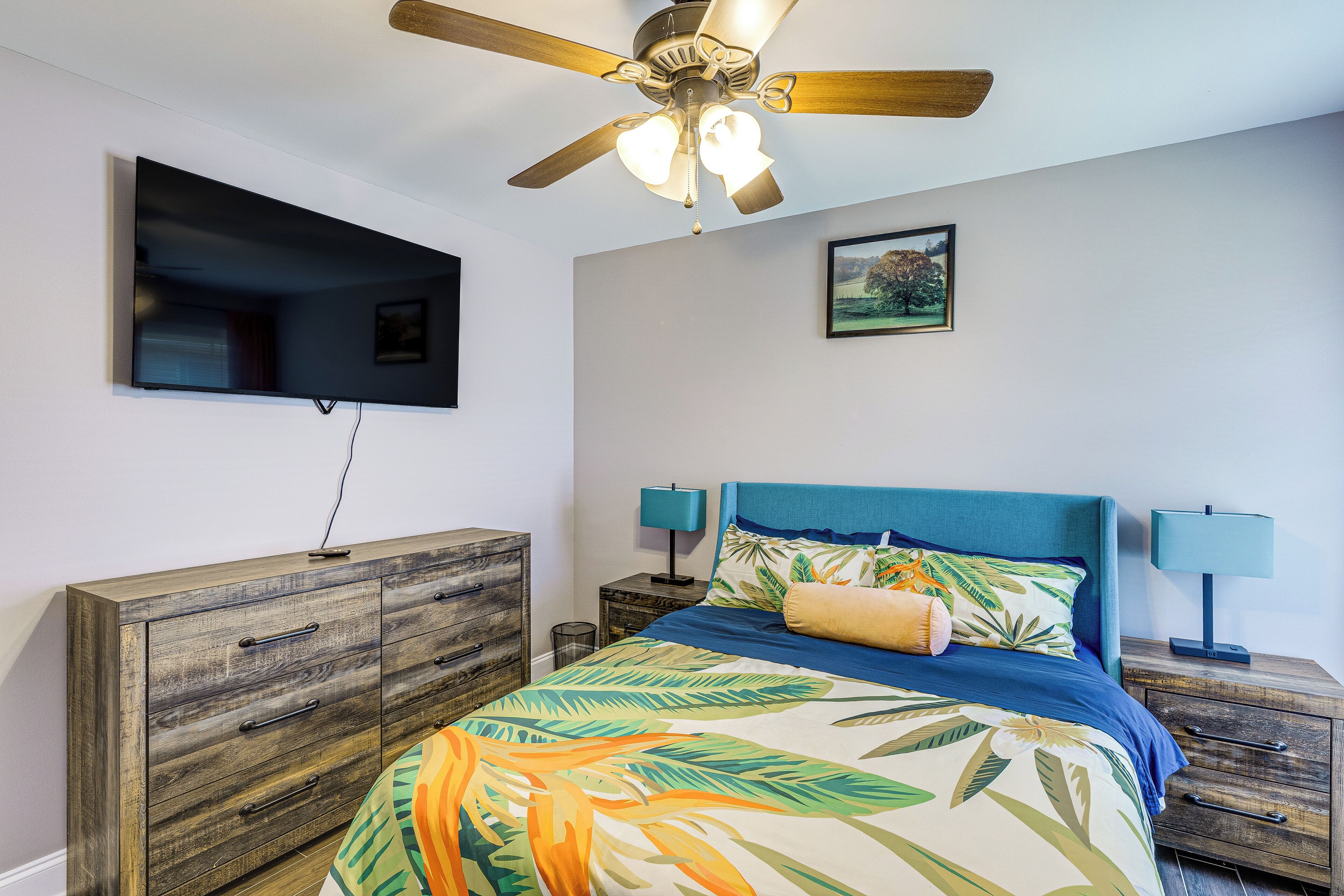 Ein Schlafzimmer mit einem Bett, einem Deckenventilator, einem an der Wand befestigten Fernseher und einem gerahmten Bild.