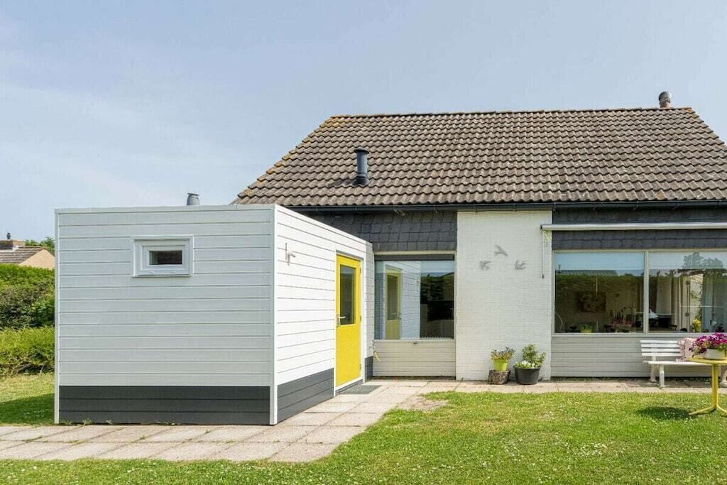 Ein modernes Haus mit Ziegeldach, ein kleiner Anbau mit gelber Tür und ein Gartenbereich mit einer Bank und Blumentöpfen.