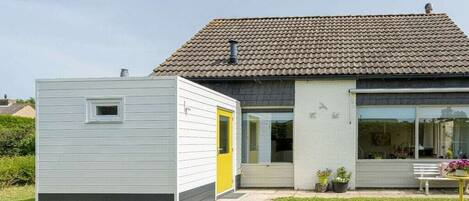 Ein modernes Haus mit Ziegeldach, ein kleiner Anbau mit gelber Tür und ein Gartenbereich mit einer Bank und Blumentöpfen.